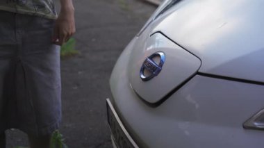 Kadın elektrikli arabayı şarj ediyor. Kapağı kaldırıyor. Şarj prizini yerleştiriyor ve uzaklaşıyor. Yüksek kalite 4K görüntü. 20 Temmuz 2020. Kyiv, Ukrayna