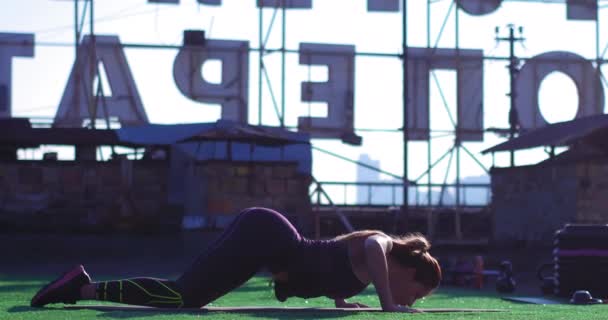 Vrouw die yoga doet op de mat op het dak van het gebouw. Het buigt zijn rug en strekt zich uit naar voren, en keert dan terug naar zijn oorspronkelijke positie. — Stockvideo