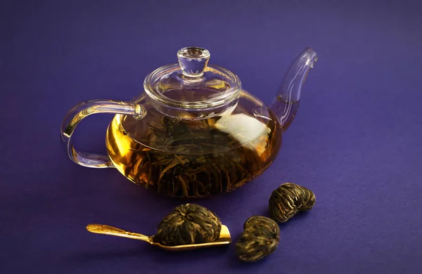 a glass teapot with a blooming tea flower on a purple background. Next to it is a Golden spoon. Close up,  selective focus