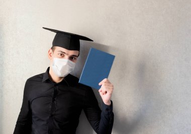 Mezuniyet şapkası ve koruyucu maskesi takmış, elinde diplomasıyla gri arka planda genç bir öğrencinin portresi. Mezuniyet 2020 konsepti