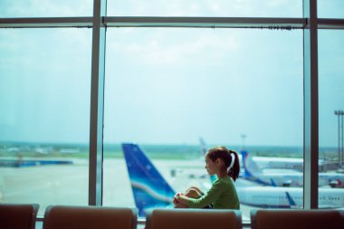 Çocuk pencerenin yanındaki havaalanında uçaklara bakıyor ve uçuş zamanını bekliyor.