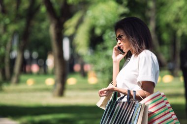 Alışveriş torbalı bir kadın alışveriş saatinden sonra şehir parkında telefonla konuşuyor.