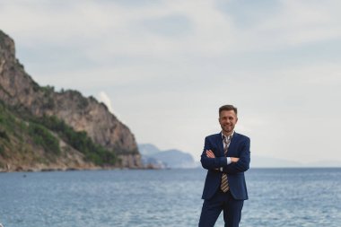 Kumsalda takım elbiseli, kravatsız bir iş adamı. Kameraya bakar ve güler, mutludur.
