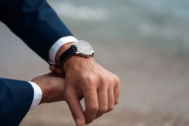 Takım elbiseli bir adam arka plandaki denizde bilek saatini kontrol ediyor.
