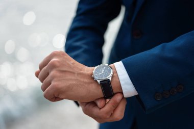 Takım elbiseli bir adam arka plandaki denizde bilek saatini kontrol ediyor.