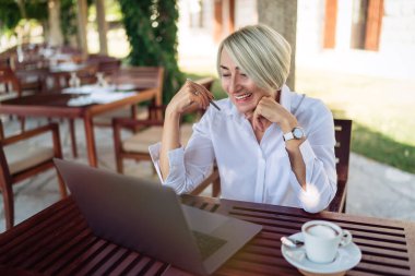 Bir kafede dizüstü bilgisayarla oturan ve kahve içen kendinden emin yaşlı bir kadın.