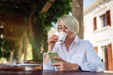 Bir kafede oturup kahve içerken cep telefonu kullanan mutlu olgun bir kadın. 