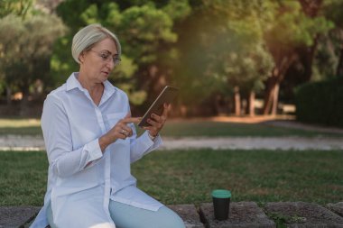Parkta dinlenirken tablet kullanan ve kahve içen olgun bir kadın.