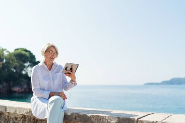 Gülümseyen olgun bir kadın arka planda tablet aracılığıyla görüntülü konuşma yapıyor deniz ve mavi gökyüzü.