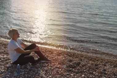 Gün batımında kumsalda koştuktan sonra oturan olgun bir kadın. Akşam koşusu