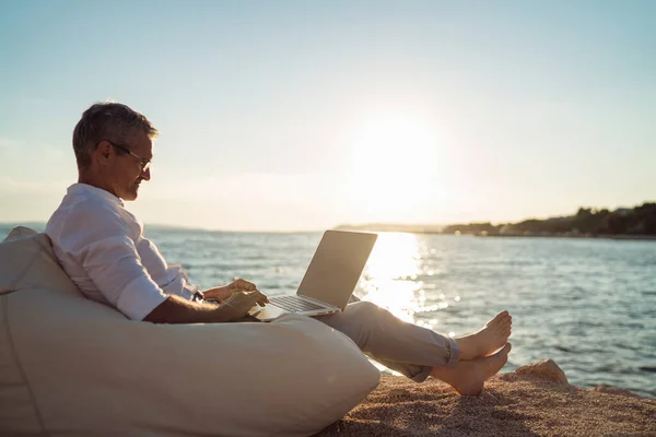 Senior man working on his laptop lying on deck chair on the beach during sunset