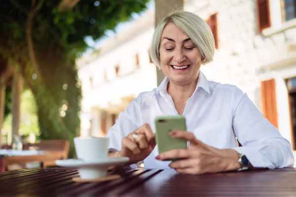 Bir kafede oturup kahve içerken cep telefonu kullanan mutlu olgun bir kadın. 