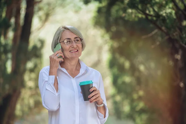 Mature woman in eyeglasses talking on mobile phone while resting at the park. Senior lady holding takeaway coffee