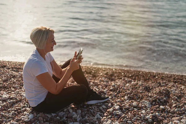 Dojrzała Kobieta Siedzi Joggingu Plaży Starsza Pani Używająca Smartfona Słuchawek — Zdjęcie stockowe