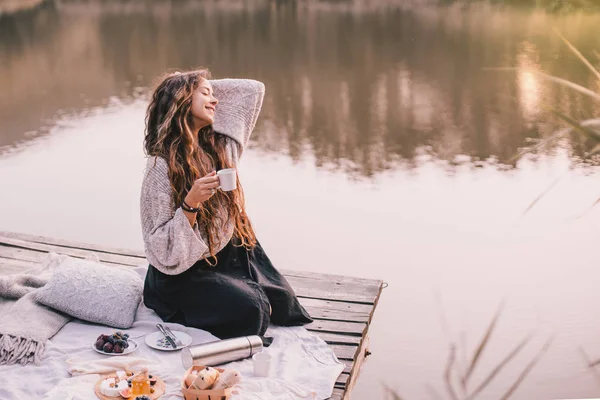 Happy woman in knitted sweater drinking tea near lake.