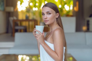 Beyaz havluya sarılı genç bir kadın kaplıcada bir fincan kahveyle oturuyor.