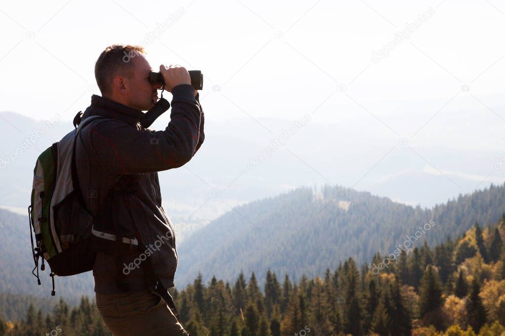 Portrait of a young man standing on a mountain hill and looking into the binoculars in the distance.