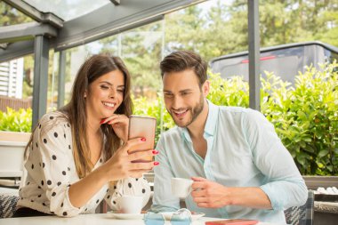 Çekici genç çift kahve içiyor ve bahçe restoranında akıllı telefon izliyor.