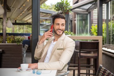 Bahçe restoranında takım elbiseli yakışıklı genç bir işadamı cep telefonuyla konuşuyor.