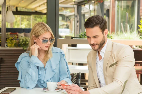 魅力的な若いカップルはコーヒーを飲み 庭のレストランでスマートフォンを見て — ストック写真