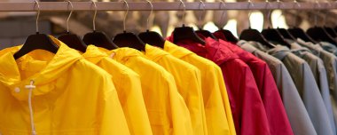Parlak renkli sonbahar yağmurlukları bir moda mağazasında asılı. Çok renkli sarı, kırmızı, gri, mavi yağmurluklu ceketler alışveriş merkezindeki giyim mağazasında askıda asılı duruyor..