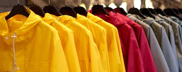 Chubasqueros de otoño de colores brillantes colgando en una tienda de moda. Primer plano de multicolor amarillo, rojo, gris, impermeables impermeables cuelgan en una fila en una percha en la tienda de ropa en el centro comercial . — Foto de Stock