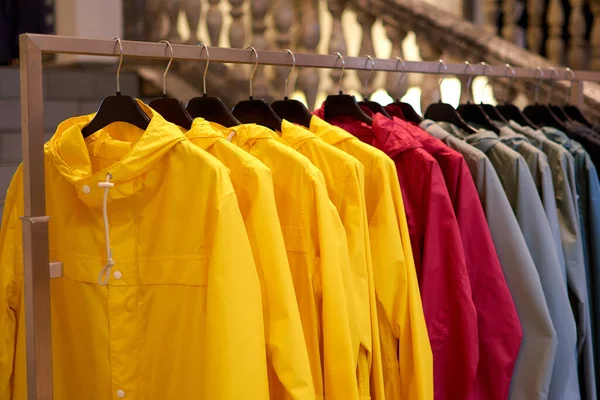 Chubasqueros de otoño de colores brillantes colgando en una tienda de moda. Primer plano de multicolor amarillo, rojo, gris, impermeables impermeables cuelgan en una fila en una percha en la tienda de ropa en el centro comercial . — Foto de Stock