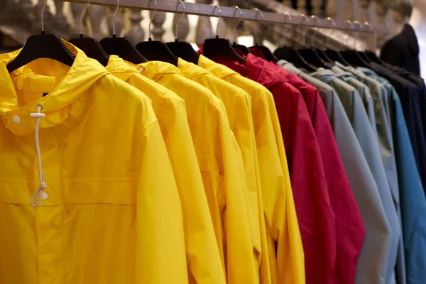 Яскраві барвисті осінні плащі висять у модному магазині. Крупним планом різнокольорові жовті, червоні, сірі, водонепроникні плащі висять поспіль на вішалці в магазині одягу в торговому центрі . Ліцензійні Стокові Фото