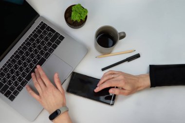 Boş defterli beyaz ofis masası, bilgisayar klavyesi, kalem, bitki ve diğer ofis malzemeleri. Fotokopi alanı olan bir kahve fincanı. Kadın eli telefonlu.