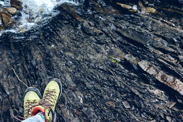 Hiking boots in outdoor action. Top View of Boot on the trail. Close-up Legs In Jeans And sport trekking shoes on rocky srones of Mountain river waterfall.