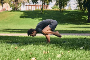 Spor giyimli genç bir adam parkta yoga yapıyor. Asana 'yı dışarıda pratik yap. Yeşil çimlerde yoga minderiyle egzersiz yapan insanlar. Karga pozu vermiş güçlü bir beyaz adam..
