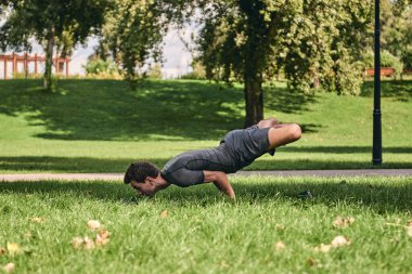 Spor giyimli genç bir adam parkta yoga yapıyor. Asana 'yı dışarıda pratik yap. Yeşil çimlerde yoga minderiyle egzersiz yapan insanlar. Nilüfer çiçeği pozu vermiş güçlü, olgun, beyaz bir adam..