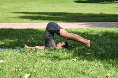 Spor giyimli genç bir adam parkta yoga yapıyor. Asana 'yı dışarıda pratik yap. Yeşil çimlerde yoga minderiyle egzersiz yapan insanlar. Plough pozu veren güçlü, olgun, beyaz adam..