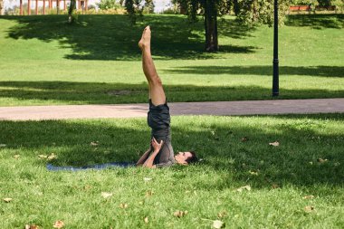 Spor giyimli genç bir adam parkta yoga yapıyor. Asana 'yı dışarıda pratik yap. Yeşil çimlerde yoga minderiyle egzersiz yapan insanlar. Omuz duruşu içinde güçlü bir beyaz adam..