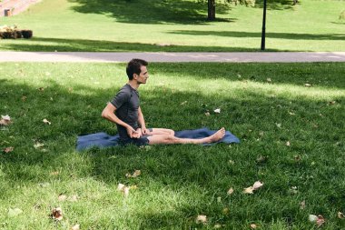 Spor giyimli genç bir adam parkta yoga yapıyor. Asana 'yı dışarıda pratik yap. Yeşil çimlerde yoga minderiyle egzersiz yapan insanlar. Fitness ve sağlıklı yaşam tarzı. Sakinlik ve meditasyon kavramı.