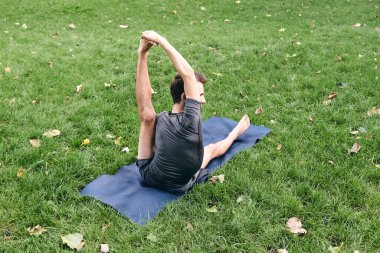 Spor giyimli genç bir adam parkta yoga yapıyor. Asana 'yı dışarıda pratik yap. İnsanlar yeşil çimlerde yoga minderiyle egzersiz yapıyorlar. Fitness ve sağlıklı yaşam tarzı. Sakinlik ve meditasyon kavramı.