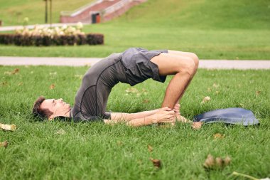 Spor giyimli genç bir adam parkta yoga yapıyor. Asana 'yı dışarıda pratik yap. Yeşil çimlerde yoga minderiyle egzersiz yapan insanlar. Güçlü, olgun, omuzlu beyaz adam Kandharasana pozu veriyor..