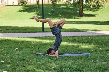 Spor giyimli genç bir adam parkta yoga yapıyor. Asana 'yı dışarıda pratik yap. Yeşil çimlerde yoga minderiyle egzersiz yapan insanlar. Nilüfer duruşu içinde zorlu amuda kalkmış güçlü beyaz adam..