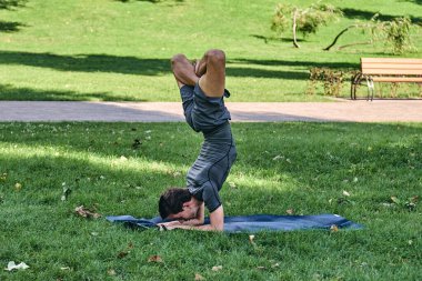 Spor giyimli genç bir adam parkta yoga yapıyor. Asana 'yı dışarıda pratik yap. Yeşil çimlerde yoga minderiyle egzersiz yapan insanlar. Nilüfer duruşu içinde zorlu amuda kalkmış güçlü beyaz adam..