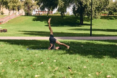 Spor giyimli genç bir adam parkta yoga yapıyor. Asana 'yı dışarıda pratik yap. Yeşil çimlerde yoga minderiyle egzersiz yapan insanlar. Omuzları ayrık, güçlü, olgun, beyaz bir adam..