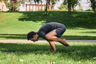 Spor giyimli genç bir adam parkta yoga yapıyor. Asana 'yı dışarıda pratik yap. Yeşil çimlerde yoga minderiyle egzersiz yapan insanlar. Karga pozu vermiş güçlü bir beyaz adam..