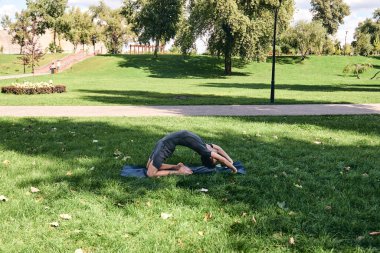 Spor giyimli genç bir adam parkta yoga yapıyor. Kapotasana asana 'yı dışarıda pratik yap. İnsanlar yeşil çimlerde yoga minderiyle egzersiz yapıyorlar. Fitness ve sağlıklı yaşam tarzı.