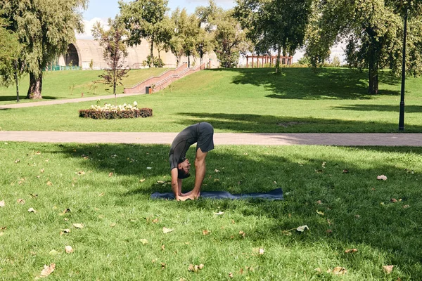 Young athletic man in sportswear doing yoga in the park. Practice standing forward bend asana pose outdoors. People exercising on green grass with yoga mat. Concept of calm and meditation.