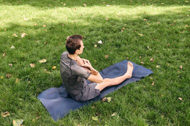 Spor giyimli genç bir adam parkta yoga yapıyor. Asana 'yı dışarıda pratik yap. Yeşil çimlerde yoga minderiyle egzersiz yapan insanlar. Fitness ve sağlıklı yaşam tarzı.