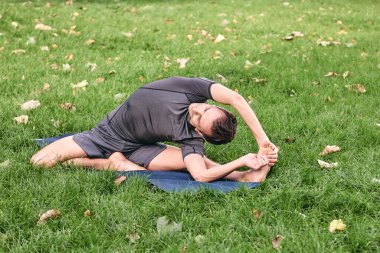 Spor giyimli genç bir adam parkta yoga yapıyor. Asana 'yı dışarıda pratik yap. Yeşil çimlerde yoga minderiyle egzersiz yapan insanlar. Parivritti Janu Sirshasana 'daki adam dizlerinin üzerine çöktü..