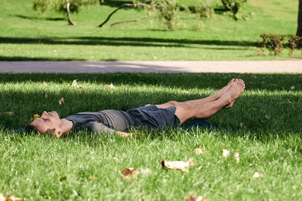 Young athletic man in sportswear doing yoga in the park. Practice asana outdoors. People exercising stretching on green grass with yoga mat. Fitness and healthy lifestyle. Concept of calm and meditation.