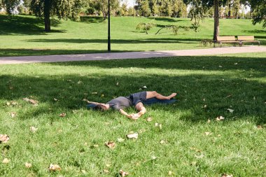 Spor giyimli genç bir adam parkta yoga yapıyor. Asana 'yı dışarıda pratik yap. İnsanlar yeşil çimlerde yoga minderiyle egzersiz yapıyorlar. Fitness ve sağlıklı yaşam tarzı. Sakinlik ve meditasyon kavramı.