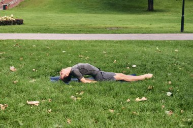 Spor giyimli genç bir adam parkta yoga yapıyor. Asana 'yı dışarıda pratik yap. İnsanlar yeşil çimlerde yoga minderiyle egzersiz yapıyorlar. Fitness ve sağlıklı yaşam tarzı. Sakinlik ve meditasyon kavramı.