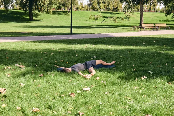 Spor giyimli genç bir adam parkta yoga yapıyor. Asana 'yı dışarıda pratik yap. İnsanlar yeşil çimlerde yoga minderiyle egzersiz yapıyorlar. Fitness ve sağlıklı yaşam tarzı. Sakinlik ve meditasyon kavramı.