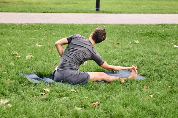 Spor giyimli genç bir adam parkta yoga yapıyor. Asana 'yı dışarıda pratik yap. Yeşil çimlerde yoga minderiyle egzersiz yapan insanlar. Parivritti Janu Sirshasana 'daki adam dizlerinin üzerine çöktü..